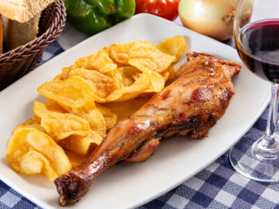 Bandeja rectangular blanca con una pata de cordero asado al horno y patatas chips crujientes junto a una copa de vino.