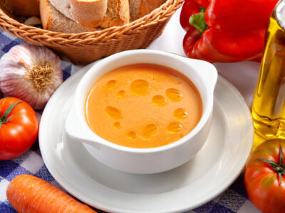 Cuenco blanco con gazpacho fresco bañado con gotas de aceite de oliva sobre plato y mantel de cuadros.
