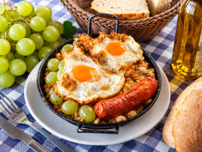 Cazuela de migas de pastor tradicionales acompañadas de huevos fritos, chorizo y uvas sobre mantel de cuadros.