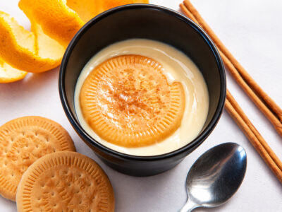 Cuenco oscuro con natillas caseras cremosas, galleta María flotando con canela, cuchara y piel de naranja.