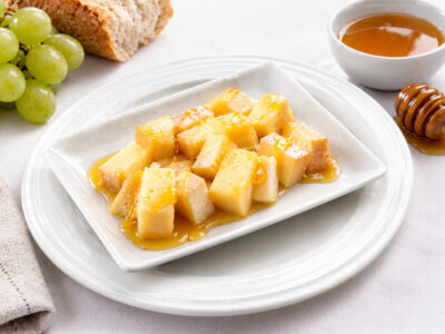 Dados de queso curado aderezados con miel en un plato blanco, acompañados de uvas verdes y un cuenco con miel.