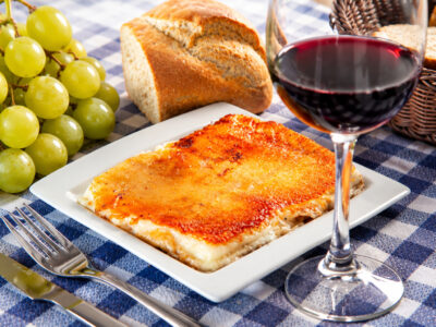Plato cuadrado con porción de queso fresco frito dorado, copa de vino tinto, racimo de uvas y pan sobre mantel de cuadros.