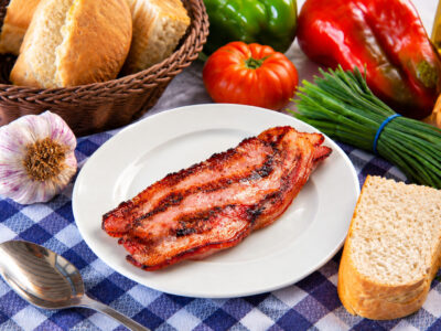 Filete de beicon o panceta a la plancha en un plato blanco, acompañado de pan, una cabeza de ajo y verduras frescas sobre un mantel de cuadros.