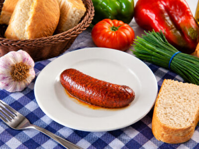 Chorizo frito en un plato blanco, acompañado de pan y verduras sobre un mantel de cuadros.