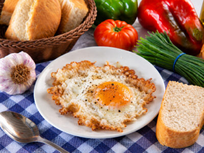 Huevo frito con la puntilla dorada en un plato blanco, acompañado de pan, una cuchara y verduras sobre un mantel de cuadros.