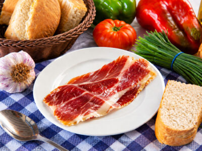 Loncha de jamón serrano en un plato blanco, acompañado de pan, una cuchara y verduras sobre un mantel de cuadros.