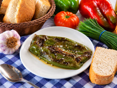 Pimiento verde asado en un plato blanco, acompañado de pan, una cuchara y verduras sobre un mantel de cuadros.