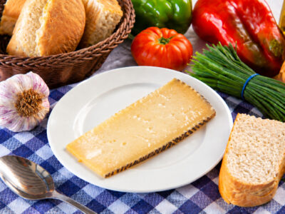 Porción de queso curado en un plato blanco, acompañado de pan, una cuchara y verduras sobre un mantel de cuadros.